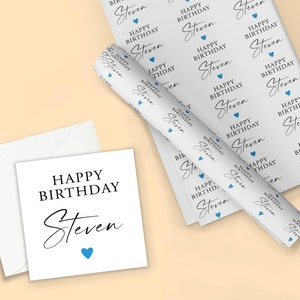 Pode incluir: Um cart&atilde;o de anivers&aacute;rio branco e um envelope a condizer com as palavras "HAPPY BIRTHDAY Steven" em escrita preta e um cora&ccedil;&atilde;o azul. Um rolo de papel de embrulho a condizer tamb&eacute;m &eacute; vis&iacute;vel, com o mesmo texto e design de cora&ccedil;&atilde;o.