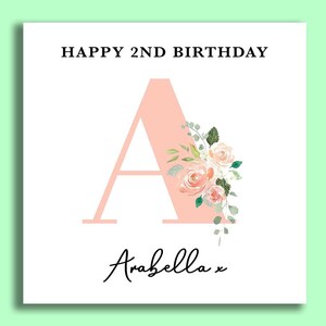 May include: A pink card with the text "HAPPY 2ND BIRTHDAY" and a large pink letter "A" decorated with flowers. The name "Arabella" is written in cursive at the bottom of the card.