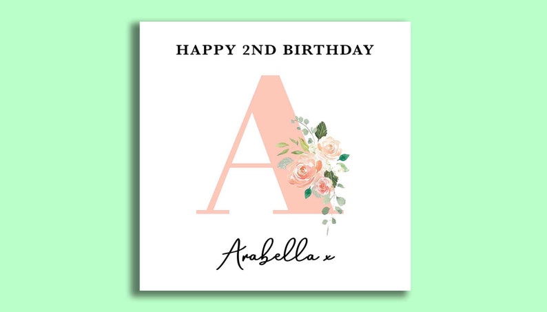 May include: A pink card with the text "HAPPY 2ND BIRTHDAY" and a large pink letter "A" decorated with flowers. The name "Arabella" is written in cursive at the bottom of the card.