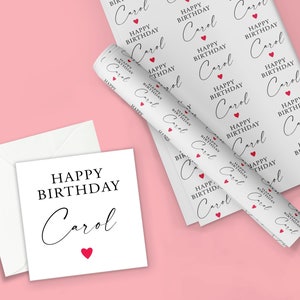 Pode incluir: Um cart&atilde;o de anivers&aacute;rio branco e um envelope a condizer com o texto "HAPPY BIRTHAY Carol" e um cora&ccedil;&atilde;o vermelho. Tamb&eacute;m &eacute; mostrado um rolo de papel de embrulho branco com o mesmo texto e design de cora&ccedil;&atilde;o.