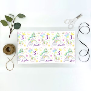Op de afbeelding: Wit inpakpapier met een herhalend patroon van pastel regenboog eenhoorns, sterren en manen. Het getal "3" en de naam "Amelie" worden herhaald in het hele ontwerp.