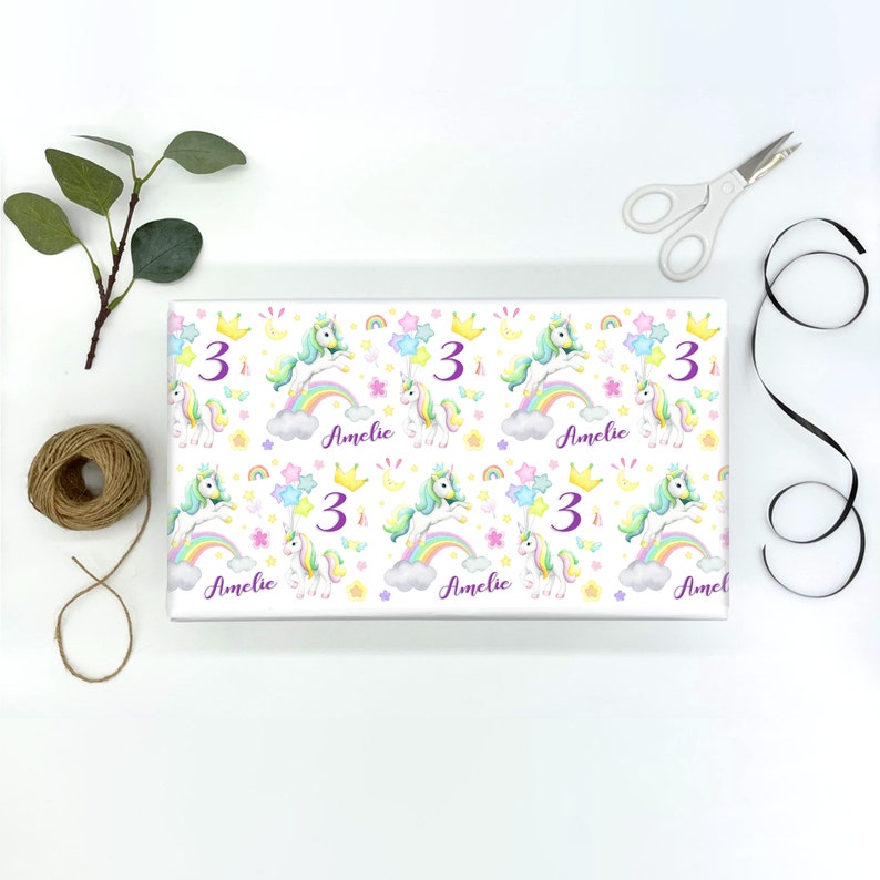 Op de afbeelding: Wit inpakpapier met een herhalend patroon van pastel regenboog eenhoorns, sterren en manen. Het getal "3" en de naam "Amelie" worden herhaald in het hele ontwerp.