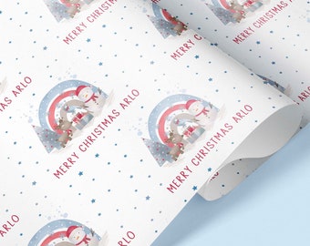 Puede incluir: Papel de regalo blanco con un patrón repetido de un muñeco de nieve, un reno y un arcoíris con el texto "Merry Christmas Arlo". El fondo está cubierto de pequeñas estrellas azules.