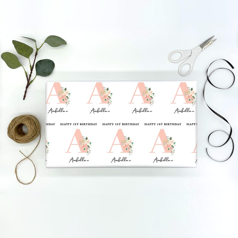 May include: Pink wrapping paper with a repeating pattern of the letter A decorated with pink flowers and the name "Ambella" written below each letter. The text "Happy 1st Birthday" is repeated on the wrapping paper.