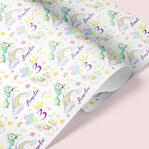 Op de afbeelding: Wit cadeaupapier met een herhalend patroon van kleurrijke eenhoorns, regenbogen, sterren en bloemen. De tekst "Amelie 3" wordt herhaald in het hele patroon.