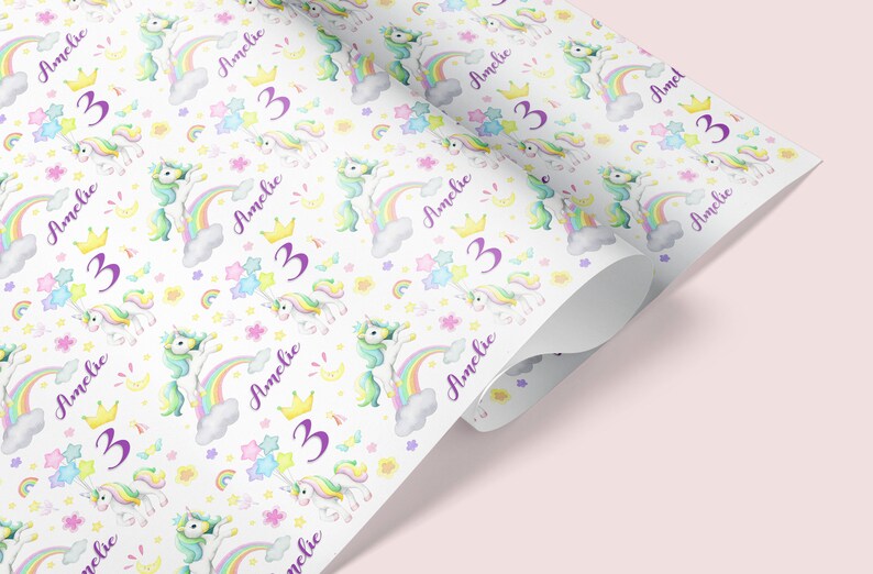 Op de afbeelding: Wit cadeaupapier met een herhalend patroon van kleurrijke eenhoorns, regenbogen, sterren en bloemen. De tekst "Amelie 3" wordt herhaald in het hele patroon.