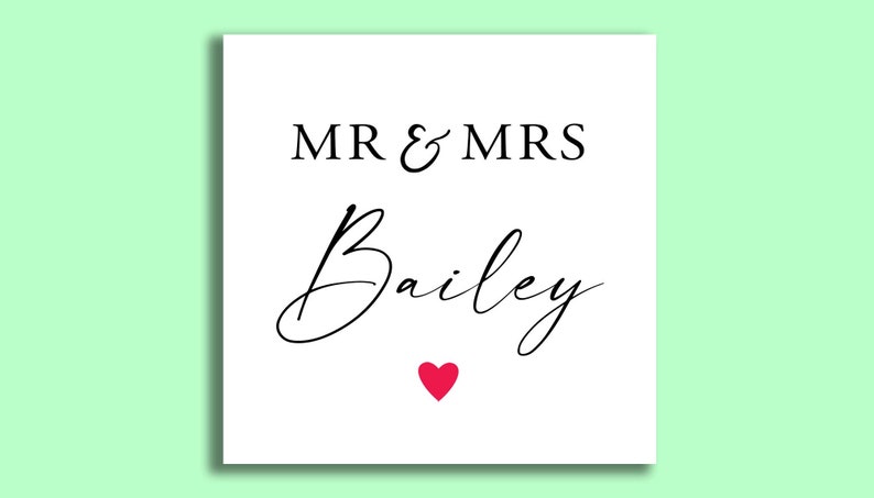 Op de afbeelding: Een witte vierkante kaart met de tekst "MR & MRS Bailey" in zwart script en een klein rood hartje onderaan. De achtergrond is lichtgroen.