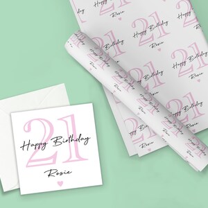 May include: A white birthday card and wrapping paper set with a pink "21 Happy Birthday Rosie" design. The wrapping paper is rolled up and laid out with the card and envelope on a light green background.
