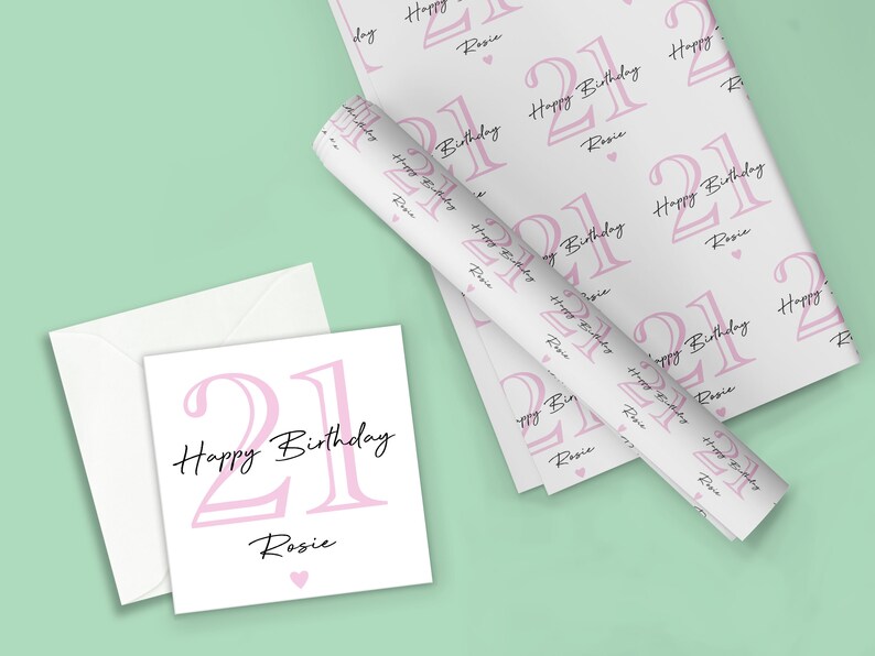 May include: A white birthday card and wrapping paper set with a pink "21 Happy Birthday Rosie" design. The wrapping paper is rolled up and laid out with the card and envelope on a light green background.