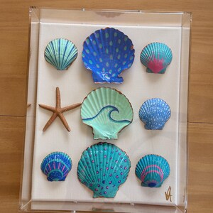 May include: A clear acrylic shadow box frame containing a collection of eight hand-painted seashells and one brown starfish arranged on a white fabric backing. The shells feature vibrant blue, green, and pink patterns, including dots and waves.