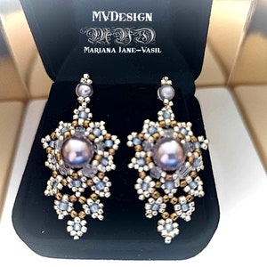 Pode incluir: Um par de brincos de contas prateadas e douradas com uma pérola grande e iridescente no centro de cada brinco. Os brincos estão pendurados em uma pequena conta prateada.