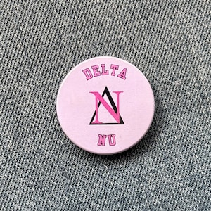 May include: A circular, pink badge featuring the words "DELTA" and "NU" in pink lettering. A black triangle and a pink "N" are in the center. The badge is set against a denim backdrop.