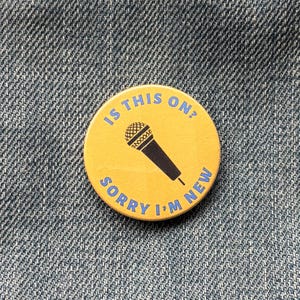 May include: A circular, yellow badge with the text "IS THIS ON?" and "SORRY I'M NEW" in blue. A black microphone graphic is in the centre. The badge is set against a denim background.
