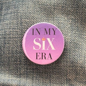 May include: A round button with a pink and purple gradient background. The button has the text "IN MY SIX ERA" in black lettering with a gold crown above the word "SIX".