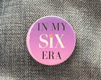May include: A round button with a pink and purple gradient background. The button has the text "IN MY SIX ERA" in black lettering with a gold crown above the word "SIX".