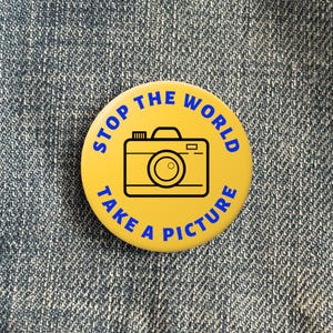 May include: Yellow button with the text "Stop the World Take a Picture" in blue letters. A black and white outline of a camera is in the centre of the button.