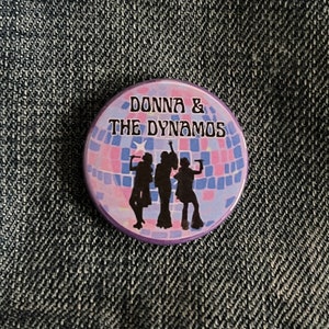 May include: A purple and pink button featuring a disco ball design and the text "Donna & The Dynamos". Three silhouettes of women are singing in front of the disco ball.