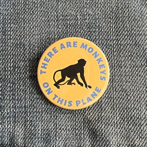 May include: A round, yellow button with blue text that reads "There are monkeys on this plane". A black silhouette of a monkey is in the centre of the button.