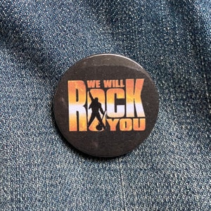 May include: A black and gold button with the text "We Will Rock You" and a silhouette of a person holding a microphone.