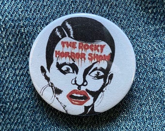 May include: A white button with a black and white illustration of a woman's face. The woman has black hair and is wearing large hoop earrings. The text "THE ROCKY HORROR SHOW" is written in red dripping letters across her forehead.