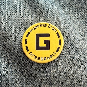 May include: A circular, yellow badge featuring the words "Pumping Iron" and "Greaseball" in black, curving around the edge. A large black "G" is in the centre. The badge is set against a denim background.