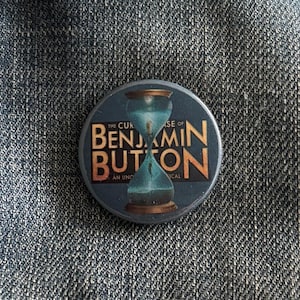 May include: A round button featuring a blue and gold hourglass design. The text "The Curious Case of Benjamin Button. An Unconventional Musical" is printed around the hourglass.