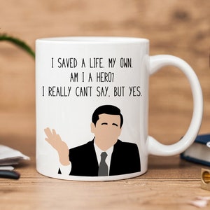 Puede incluir: Taza de café de cerámica blanca con una ilustración en blanco y negro de un hombre con el texto "Salvé una vida. La mía. ¿Soy un héroe? Realmente no puedo decirlo, pero sí."