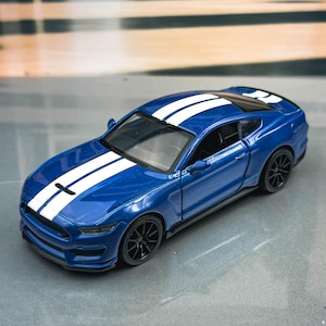 May include: A blue toy car with white racing stripes on the hood and roof. The miniature car has black wheels and a detailed design, resembling a sports car. The car is set against a blurred background.