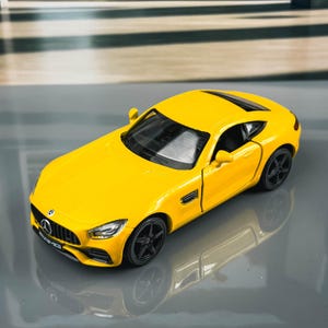 May include: A bright yellow die-cast model car with black wheels. The miniature sports car has a detailed front grille and headlights. The car is a miniature replica of a sports car.
