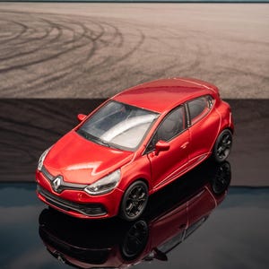Può includere: Un'auto modello Renault Clio rossa con ruote nere si riflette su una superficie nera lucida. L'auto è ambientata sullo sfondo di un circuito con segni di pneumatici, montagne e cielo blu.