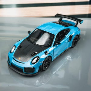 May include: A light blue toy car with a black bonnet and spoiler. The miniature vehicle has black wheels and headlights. The car is a model of a sports car.