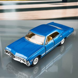 May include: A blue die-cast model car with white-walled tyres and a chrome grill. The miniature vehicle has a tan interior and is set against a blurred background of trees and architecture. The car is a classic vintage model.