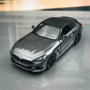 Op de afbeelding: Een grijs model van een cabriolet met een zwart dak. De miniatuurauto heeft zwarte wielen en een gedetailleerde grille. De achtergrond toont een vage buitenscène met bomen en een gebouw.