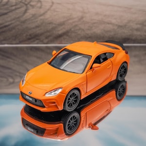 Peut inclure: Une voiture de sport miniature orange vif en métal moulé sous pression, dotée d'une carrosserie coupé profilée, de roues noires à rayons multiples, d'un aileron arrière noir et de phares clairs. Le véhicule est posé sur une surface réfléchissante.