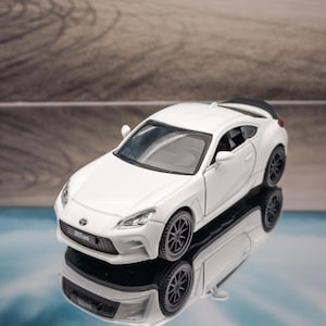 May include: A white Toyota GR86 scale model car featuring a sleek coupe body, black multi-spoke wheels, a black rear spoiler, and a detailed front grille with the Toyota emblem centered on the hood, reflecting on a smooth, dark surface.