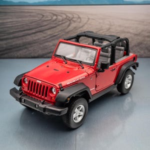 Könnte beinhalten: Ein rotes Jeep Rubicon Modell mit schwarzen Akzenten, einem schwarzen Überrollkäfig und silbernen Rädern. Das Spielzeugauto steht auf einer reflektierenden Oberfläche mit verschwommenem Hintergrund von Straße und Bergen.