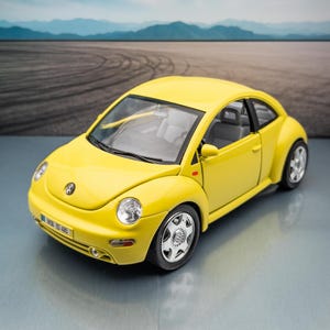 Puede incluir: Un coche de juguete Volkswagen Beetle amarillo con ruedas plateadas. El coche tiene un interior negro y una matrícula que dice "WOB-BD 485". El fondo presenta un paisaje borroso y un cielo azul.