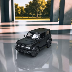 May include: A black die-cast model of a Ford Bronco SUV. The model features detailed headlights, a front grille with the "BRONCO" logo, and black wheels. The vehicle is a two-door model, and the overall design is a miniature replica of the full-size SUV.