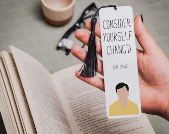 May include: White bookmark with black text that reads "Consider yourself Chang'd - Ben Chang". A cartoon image of Ben Chang from the TV show Community is at the bottom of the bookmark. The bookmark has a black tassel.