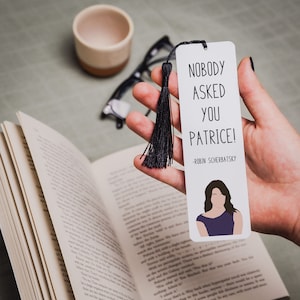 May include: A white bookmark with black text that reads "Nobody asked you Patrice! -Robin Scherbatsky". A cartoon illustration of a woman with long dark hair wearing a purple top is at the bottom of the bookmark.