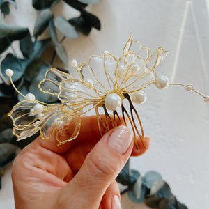 Op de afbeelding: Een goudkleurige haar kam met een delicaat bloemmotief. De kam is versierd met transparante, bloemblad-vormige elementen en imitatieparels van verschillende groottes. De tanden van de kam zijn zichtbaar, en het ontwerp is elegant, geschikt voor bruiden of formele gelegenheden.