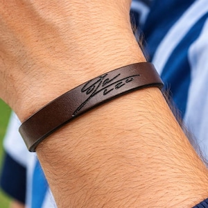 May include: A dark brown leather bracelet featuring a smooth, flat band design. The surface of the leather is embossed with a black, stylized cursive signature that reads "Sia Lee" in a flowing, handwritten script style across the center.