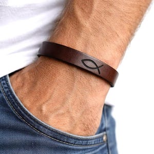 May include: A dark brown leather bracelet featuring a debossed fish symbol. The bracelet is worn on a wrist, with a white shirt and blue jeans in the background. The bracelet has a minimalist design.