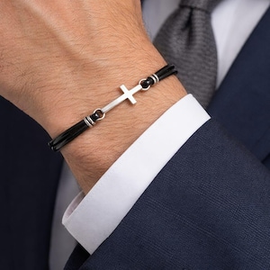 Può includere: Un braccialetto con croce argentata e cordino nero. La croce è il punto focale del braccialetto, e il design è semplice ed elegante. Il braccialetto è indossato al polso, e lo sfondo include un abito e una cravatta.