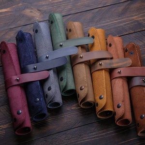 Puede incluir: Un conjunto de nueve estuches para lápices de cuero en varios colores, incluyendo rojo, azul, gris, verde, marrón y naranja. Cada estuche tiene una correa de cuero con una hebilla de metal.