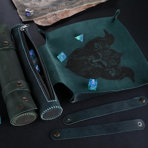 May include: A green leather dice rolling tray with a black and white illustration of a creature with horns. The tray has two matching green leather dice rolling tubes with a zipper closure. There are several blue and green dice on the tray and tubes.