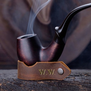 Könnte beinhalten: Eine dunkelbraune Holzpfeife mit einem Lederhalter. Der Halter hat die Initialen "W.W." geprägt. Rauch steigt aus der Pfeife auf.