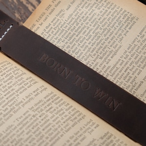 Pode incluir: Um marcador de p&aacute;gina de couro castanho com o texto "N.L." e "BORN TO WIN" gravado em relevo. O marcador de p&aacute;gina est&aacute; sobre um livro.