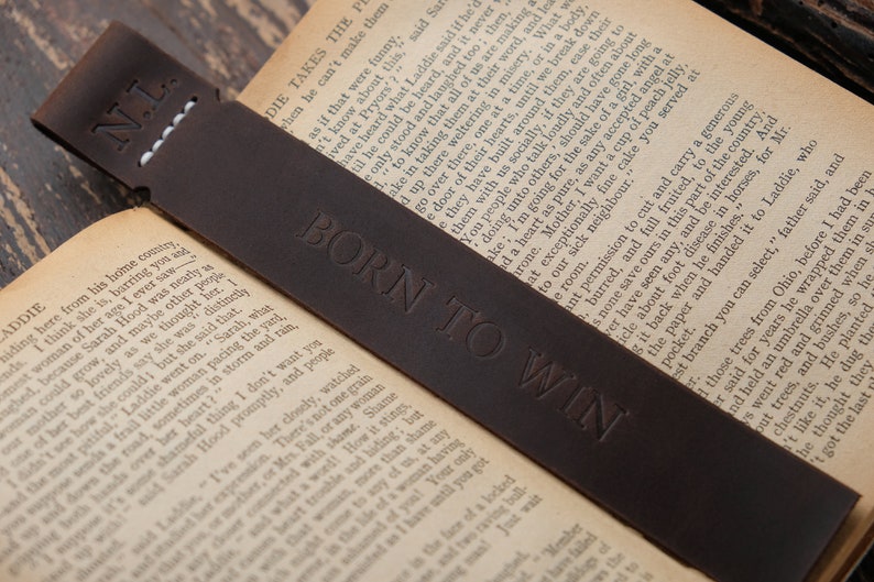 Pode incluir: Um marcador de p&aacute;gina de couro castanho com o texto "N.L." e "BORN TO WIN" gravado em relevo. O marcador de p&aacute;gina est&aacute; sobre um livro.