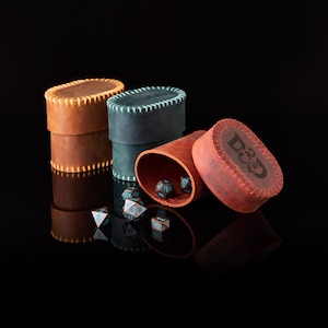 May include: Three oval leather dice containers in tan, dark blue, and reddish-brown. The reddish-brown container is open, revealing polyhedral dice with blue numbers. The lid of the reddish-brown container features the D&D logo and text.